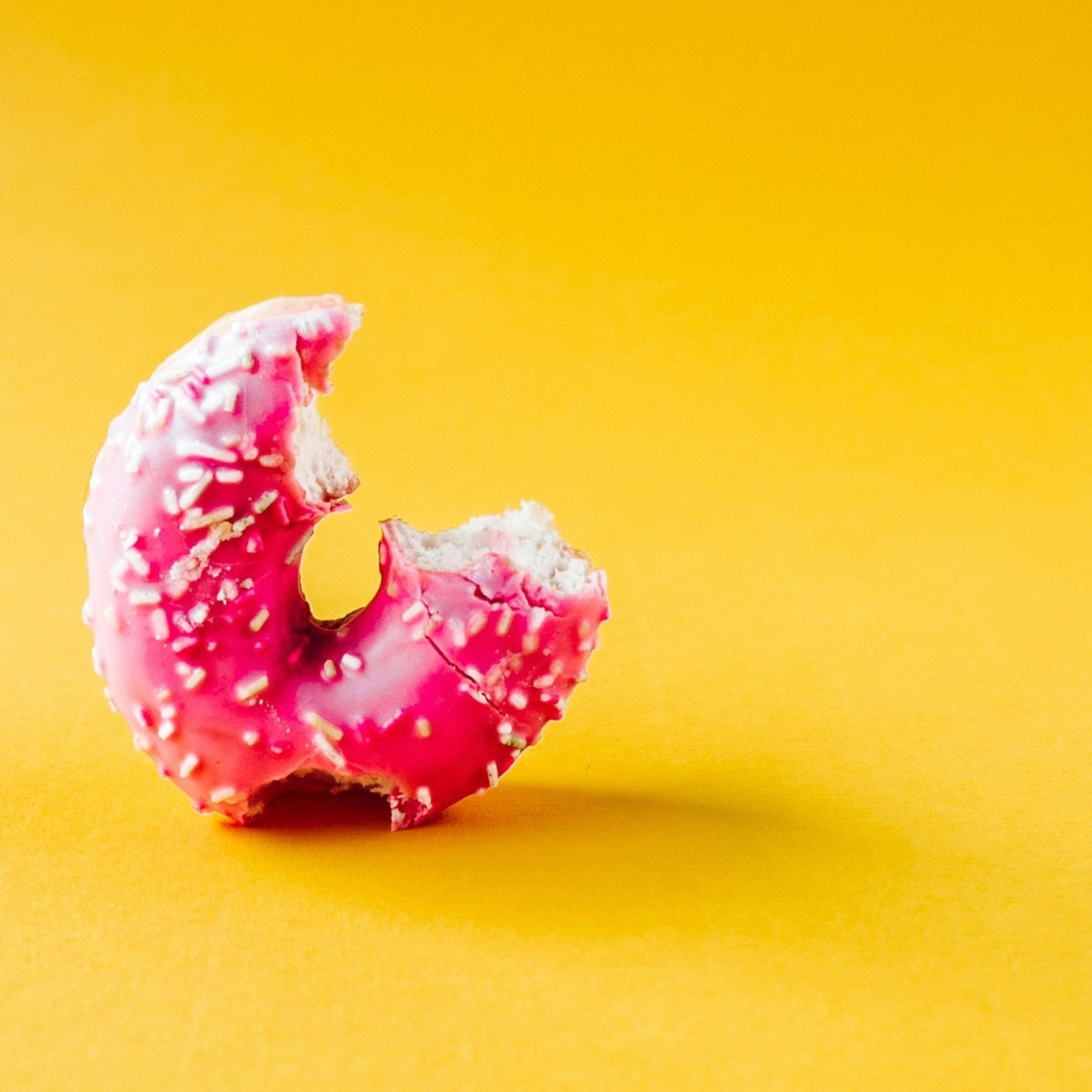 Pink ring doughnut with sprinkles on a yellow background. There are two bites missing.