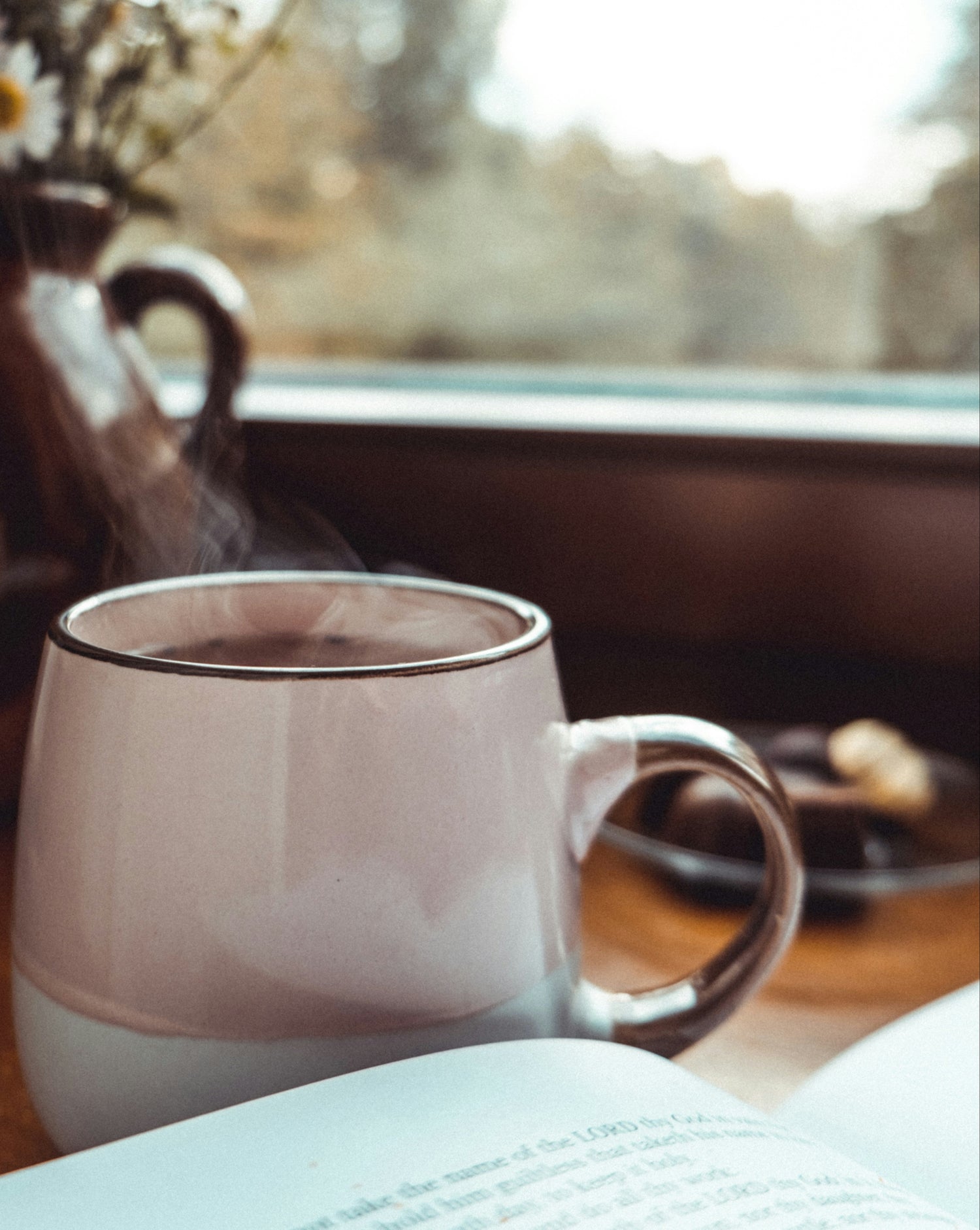 Steaming mug of coffee next to an open book with a window and flowers in the background